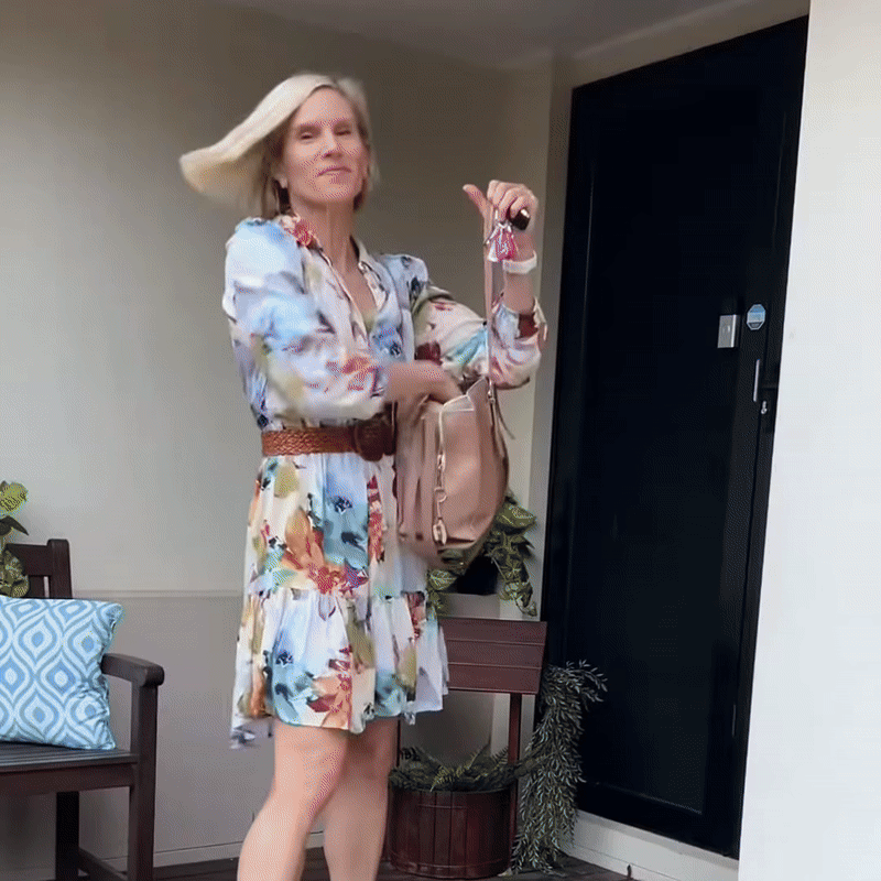 Vrouw in bloemetjesjurk met schouderlang blond haar dat wappert terwijl ze met sleutels en tas voor een deur staat. 8