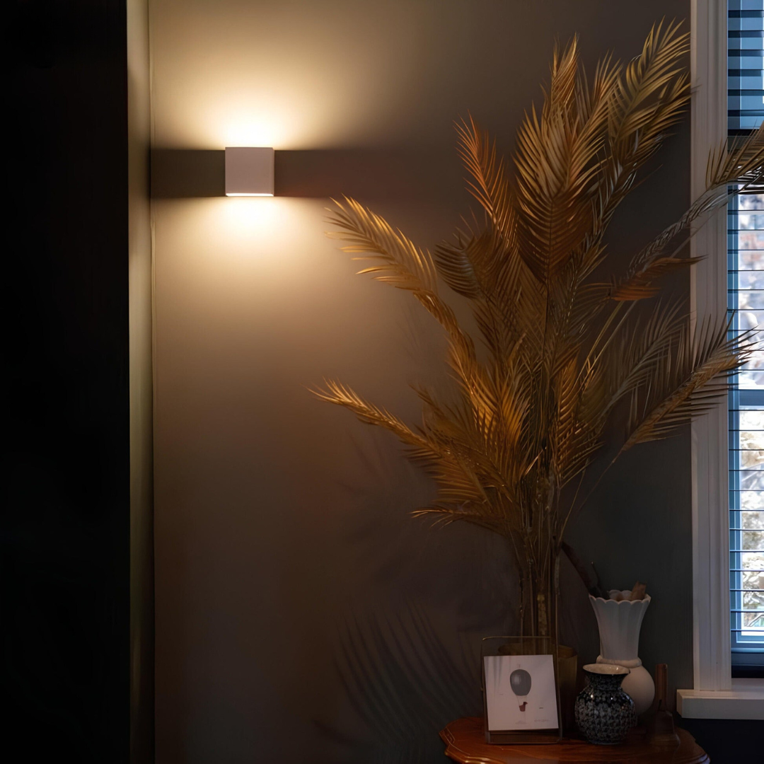 Witte kubuslamp op een donkere muur naast een grote bos gouden palmbladeren op een tafeltje. 4