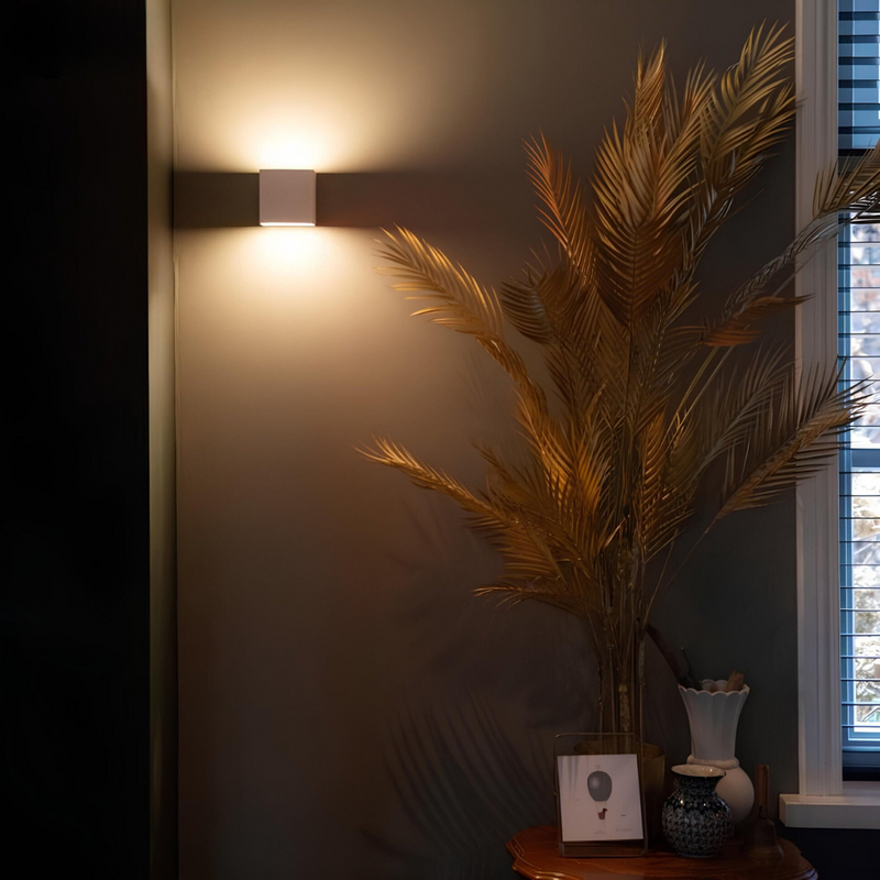 Witte kubuslamp op een donkere muur naast een grote bos gouden palmbladeren op een tafeltje. 4