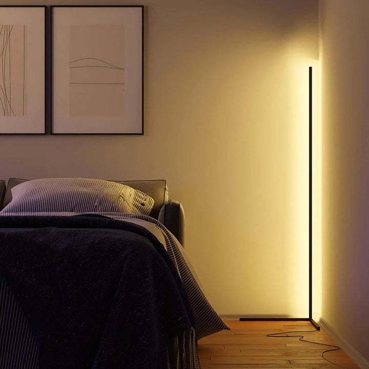 Lamp in de hoek van een slaapkamer die de muur warm verlicht, naast een bed en ingelijste prints 4