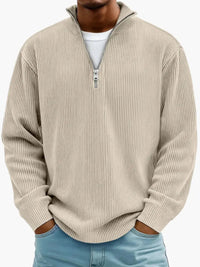 De geribbelde trui met ritskraag in beige, gedragen door een model over een wit shirt en met een lichtblauwe spijkerbroek. 4