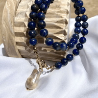 Dubbel gelegde blauwe kralenketting met parelhanger op wit doek, tegen een houten ronde sculptuur 7