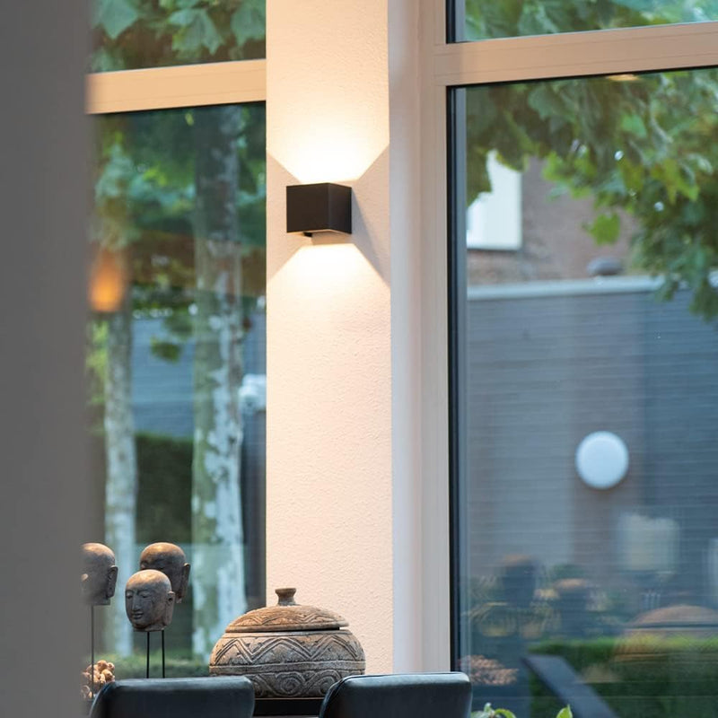 Zwarte wandlamp op een lichte pilaar tussen grote ramen, met decoratieve hoofdjes en een houten pot op de voorgrond. 8