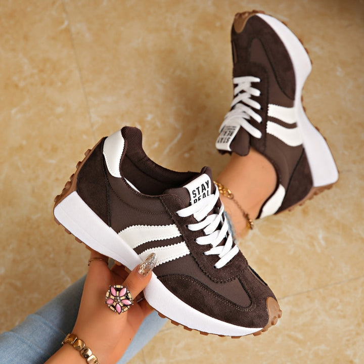 Bruine suède sneakers met witte strepen, één in de hand gehouden en één aan de voet 3