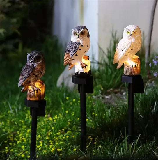 Drie uilen in bruin, bruinwit en wit staan 's avonds verlicht op pinnen in het gras met kleine gele bloemetjes. 8