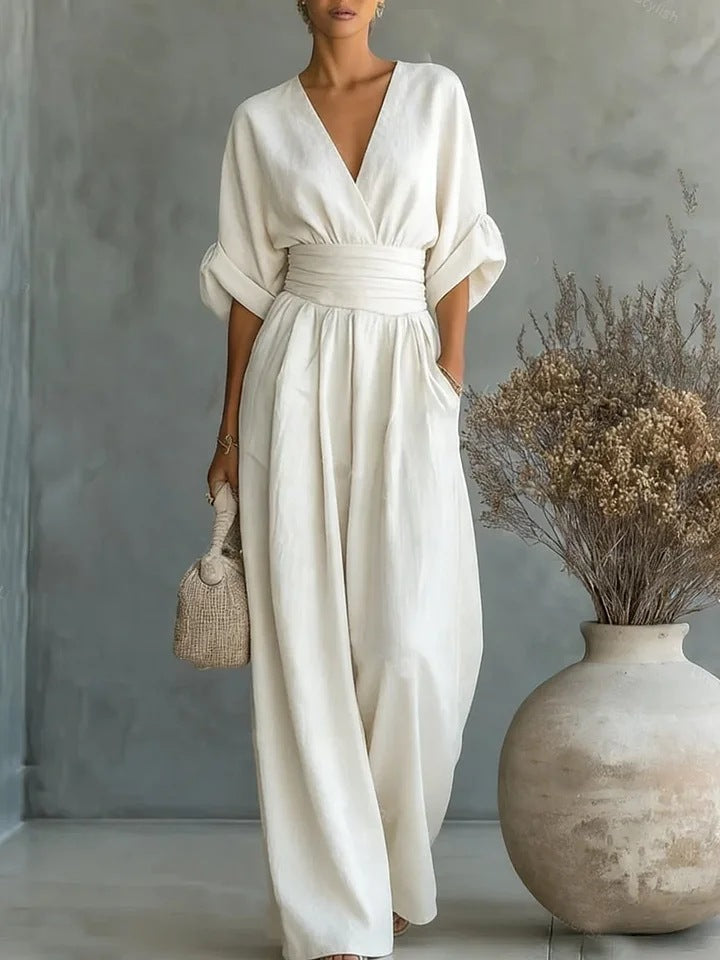 Vrouw in crèmewitte jumpsuit met brede ceintuur en halflange mouwen, een rieten tasje in de hand, naast een grote aardewerken vaas. 1