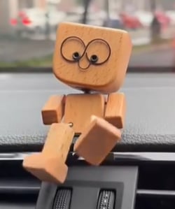 Houten figuurtje met grote ogen en een kleine mond zit op een autodashboard, met een raam op de achtergrond. 6