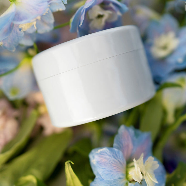 Witte ronde inzet ligt tussen blauwe en witte bloemen en groene bladeren, met de naad van het deksel zichtbaar. 9