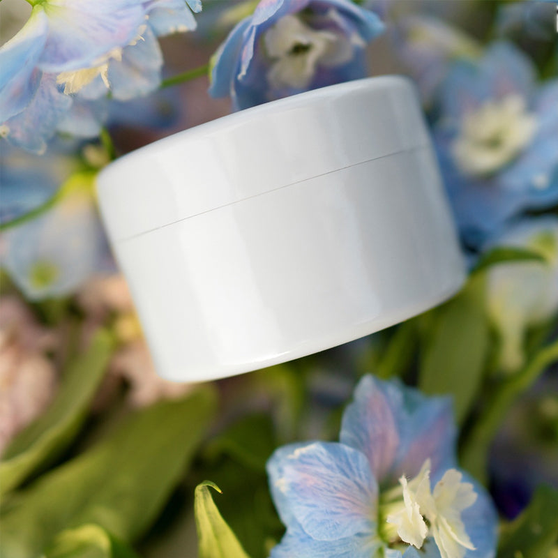 Witte ronde inzet ligt tussen blauwe en witte bloemen en groene bladeren, met de naad van het deksel zichtbaar. 15