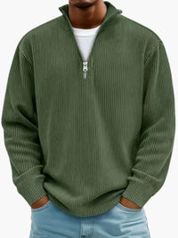 De geribbelde trui met kwartrits in legergroen, gedragen door een model met wit shirt eronder en handen in de zakken. 8