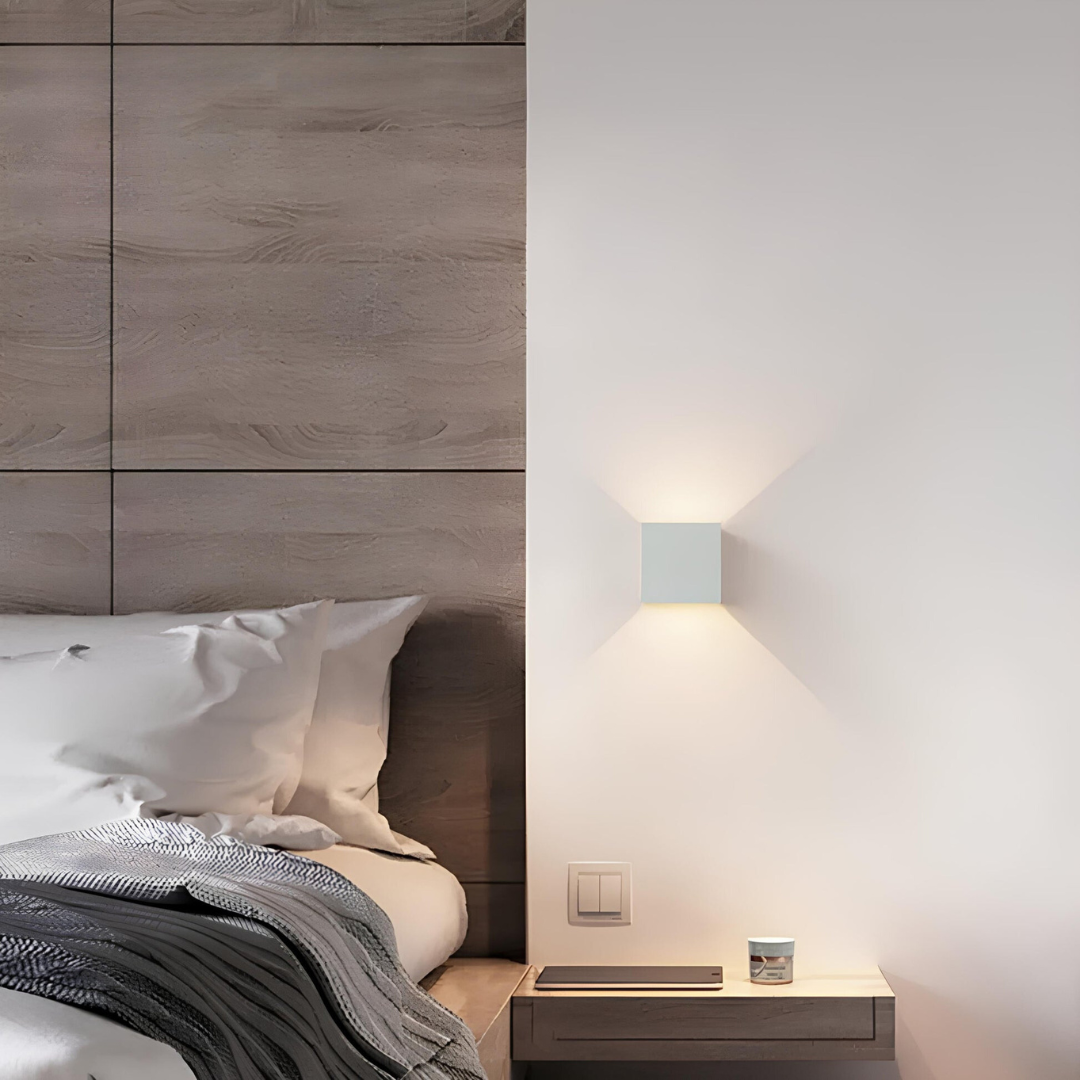 Witte kubuslamp aan de muur naast een bed met houten hoofdbord en een kleine plank als nachtkastje. 6