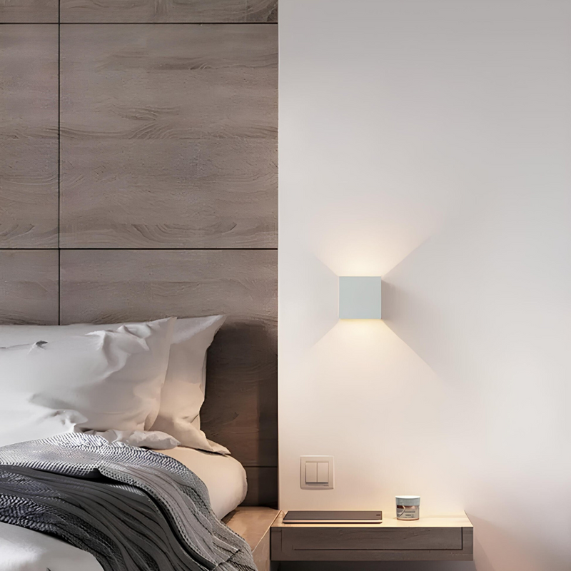 Witte kubuslamp aan de muur naast een bed met houten hoofdbord en een kleine plank als nachtkastje. 6