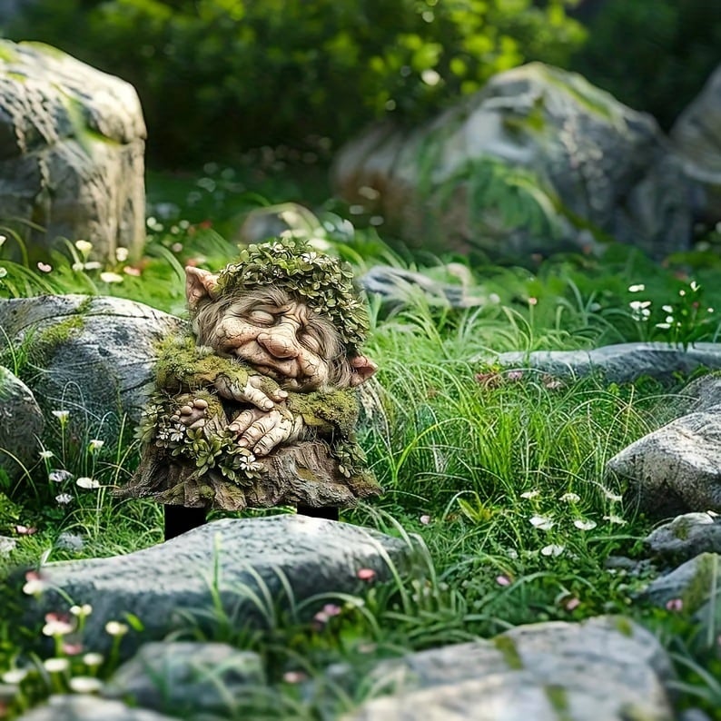 Tuinprikker van de slapende bemoste trol met bladkrans, tussen rotsen en gras met kleine witte bloemetjes. 16