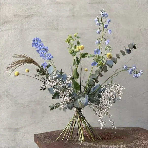 Los gebonden boeket met blauwe bloemen, gele bolletjes en eucalyptus, aan de stelen bijeengehouden en tegen een grijze muur getoond. 12