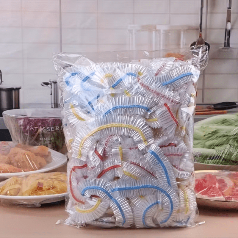Grote doorzichtige zak vol gevouwen elastische deksels met blauwe, gele en rode randen, op een keukenblad naast afgedekte schalen met eten. 1