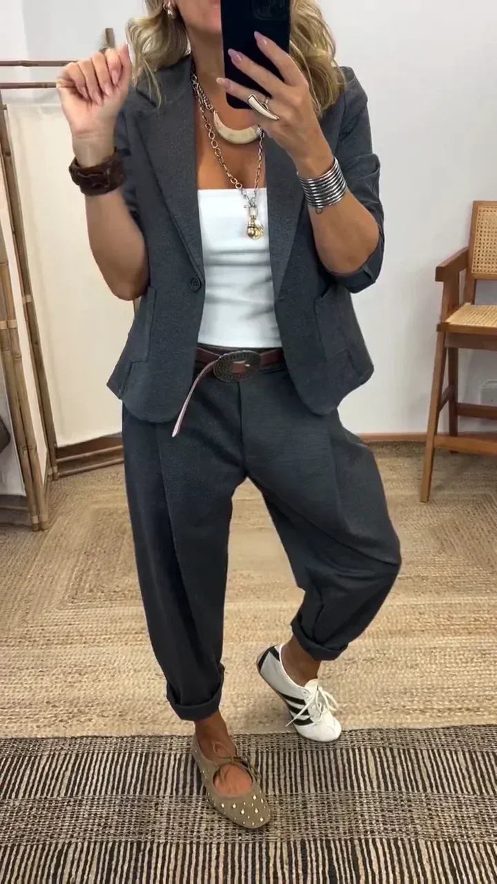 Vrouw in donkergrijs kostuum met blazer met één knoop en ruime broek met omgeslagen pijpen, gedragen met wit topje en bruine riem. 1