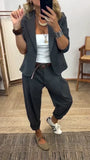 Vrouw in donkergrijs kostuum met blazer met één knoop en ruime broek met omgeslagen pijpen, gedragen met wit topje en bruine riem. 1