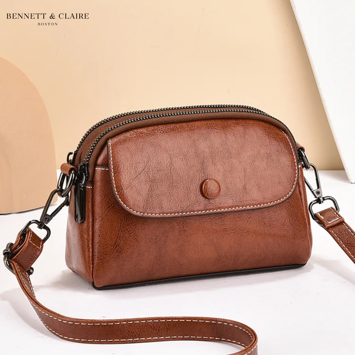 Cognacbruine schoudertas met klepje, drukknoop, witte stikselrand en dubbele rits, schuin van voren gefotografeerd met de band op de grond. 1