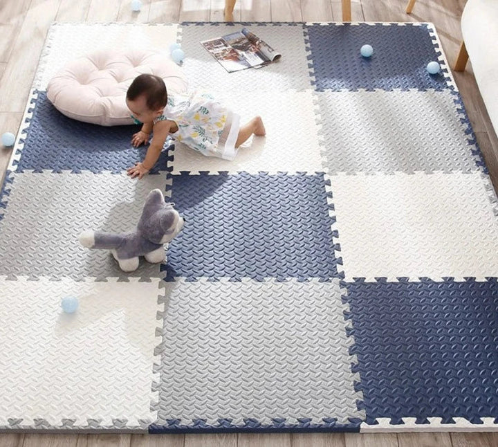 Baby kruipt over een speelmat van blauwe, grijze en witte puzzeltegels, met knuffelhond en kleine ballen erbij. 2