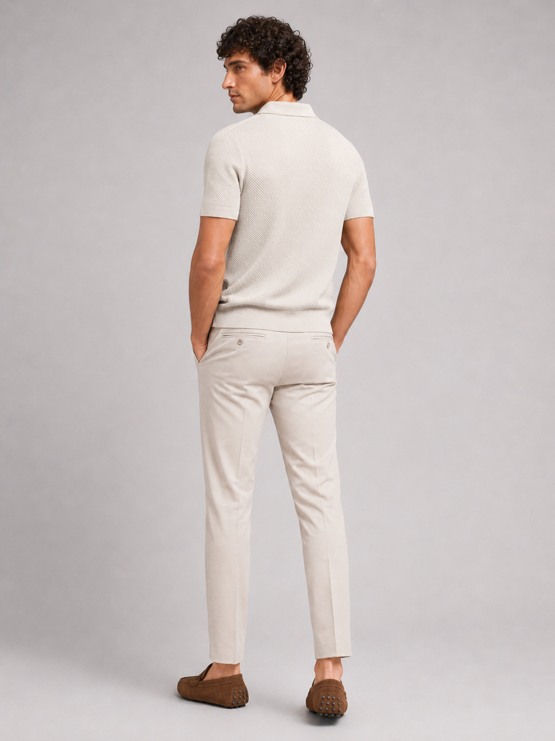 Achterkant van het beige poloshirt, model kijkt over zijn schouder, met crèmekleurige broek en bruine loafers 19