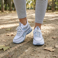 Gedragen door een vrouw in grijze legging op een bospad met herfstbladeren: witte sneakers met lichtblauwe zool, voorkant. 2