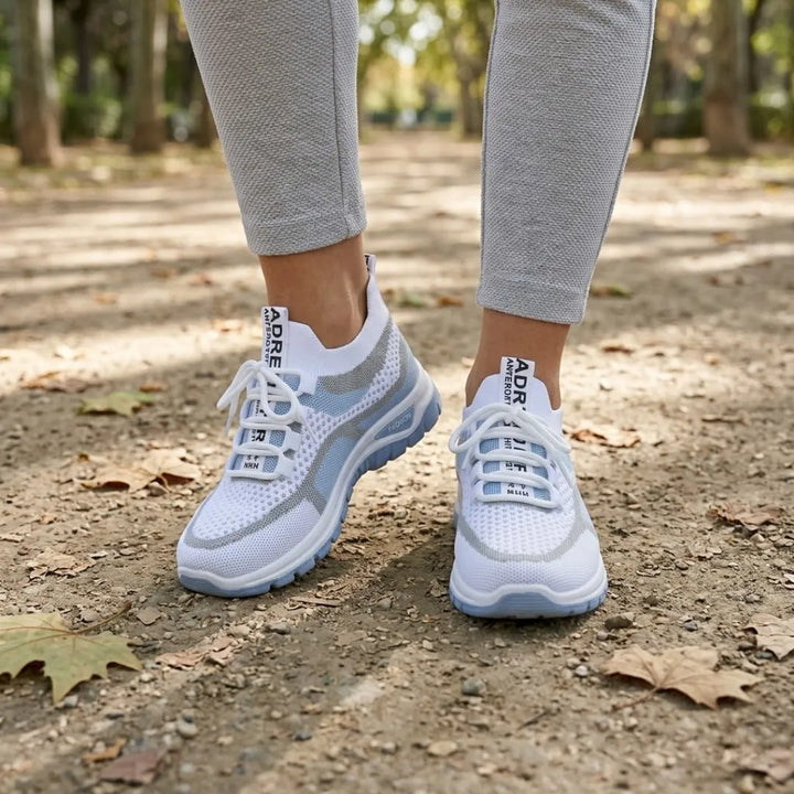 Gedragen door een vrouw in grijze legging op een bospad met herfstbladeren: witte sneakers met lichtblauwe zool, voorkant. 2