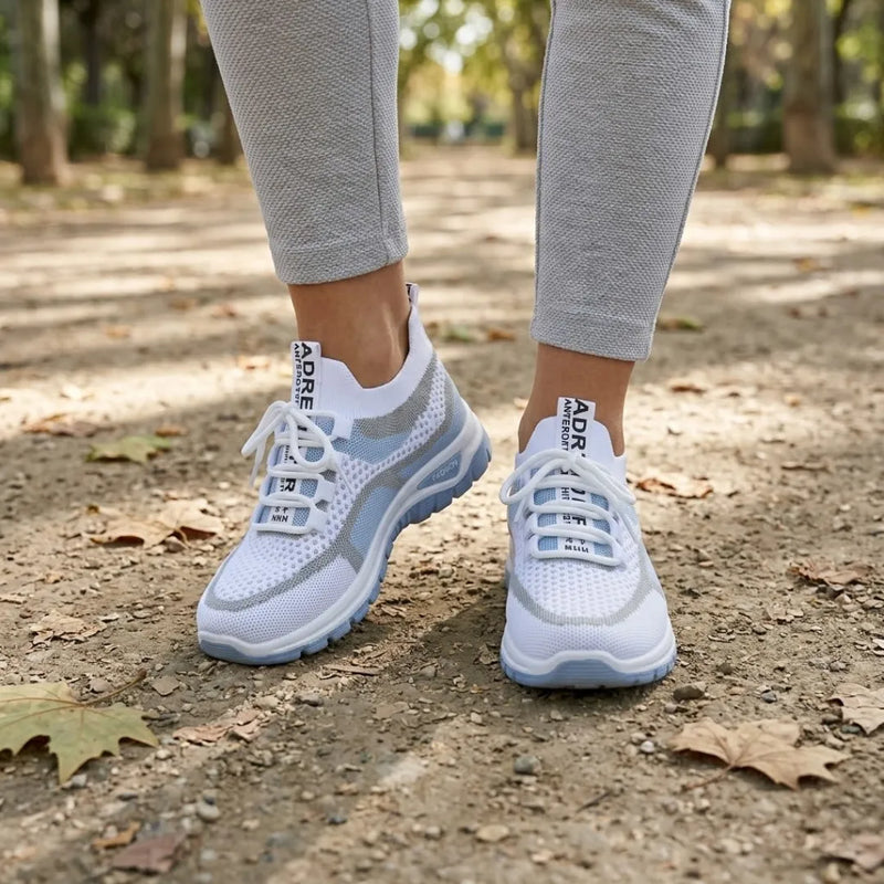 Gedragen door een vrouw in grijze legging op een bospad met herfstbladeren: witte sneakers met lichtblauwe zool, voorkant. 2