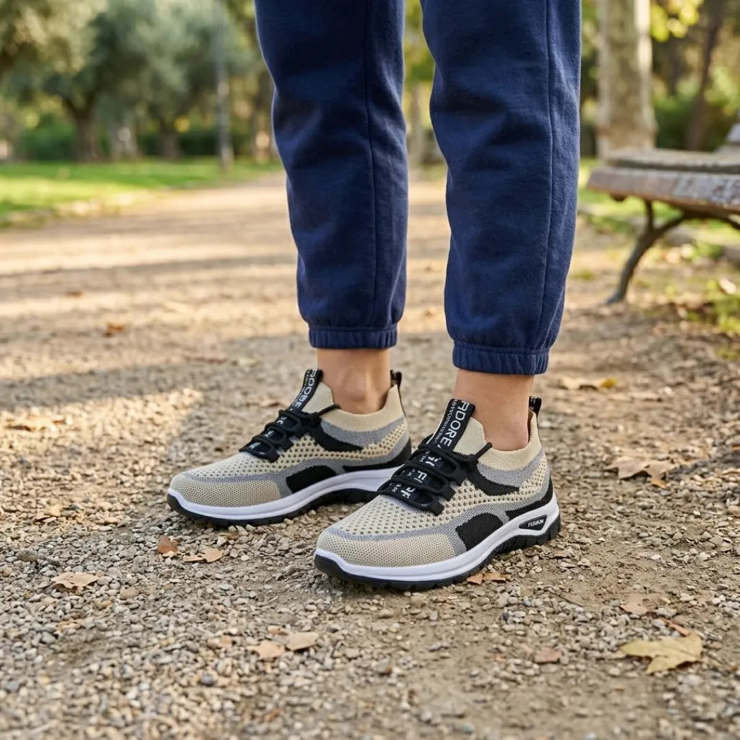Voeten van een persoon in een blauwe joggingbroek met beige en zwarte gaas sneakers, staand op een zandpad in een park. 38