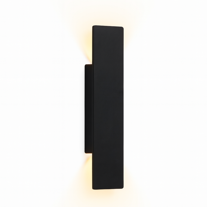 Zwarte rechthoekige wandlamp schuin van opzij op een witte achtergrond, met warm licht dat boven en onder langs de montageplaat schijnt. 1