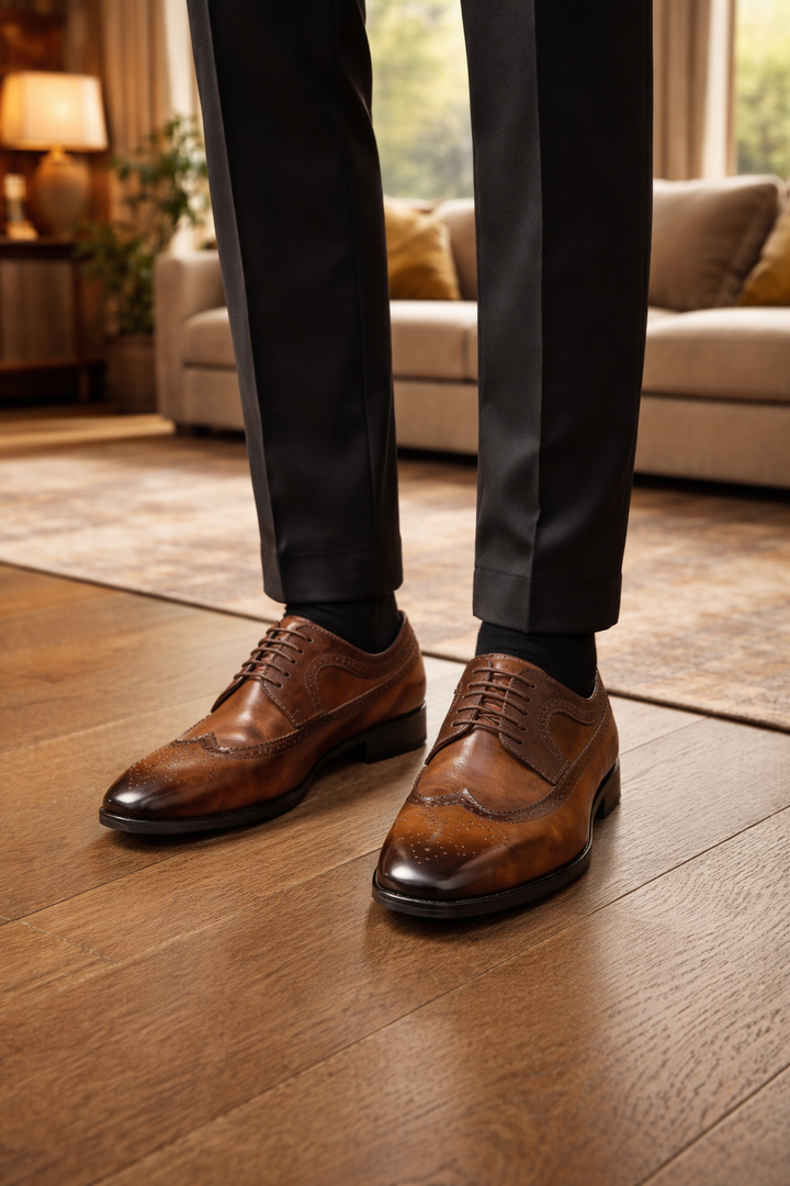 Bruine brogue veterschoenen gedragen met een donkere pantalon en zwarte sokken, op een houten vloer in een woonkamer. 1