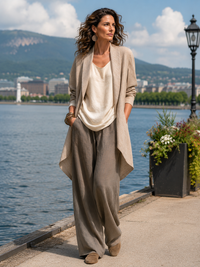 Model draagt de driedelige set buiten langs het water, met beige open jack, crèmekleurige top en taupe wijde broek. 7