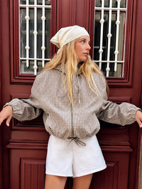 Model draagt de geruite jas open armen tegen een rode deur, met witte short en een lichte hoofddoek. 1