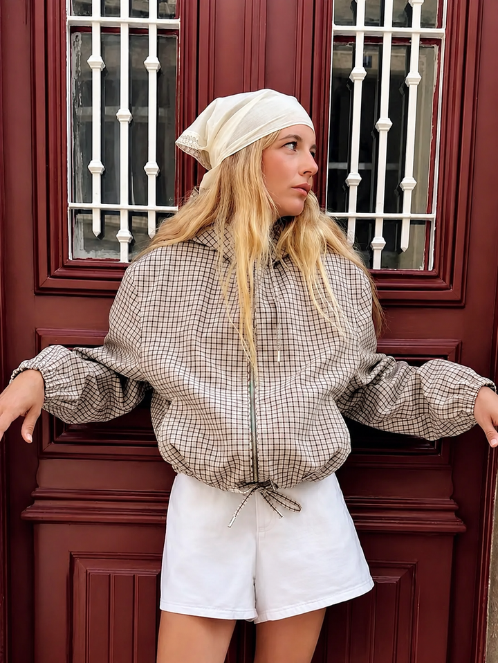 Model draagt de geruite jas open armen tegen een rode deur, met witte short en een lichte hoofddoek. 1