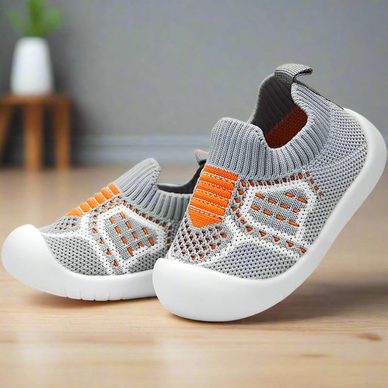 Grijze gebreide babyschoentjes met oranje streepdetails, geribbelde boord en witte zool, schuin geplaatst op een houten vloer. 16