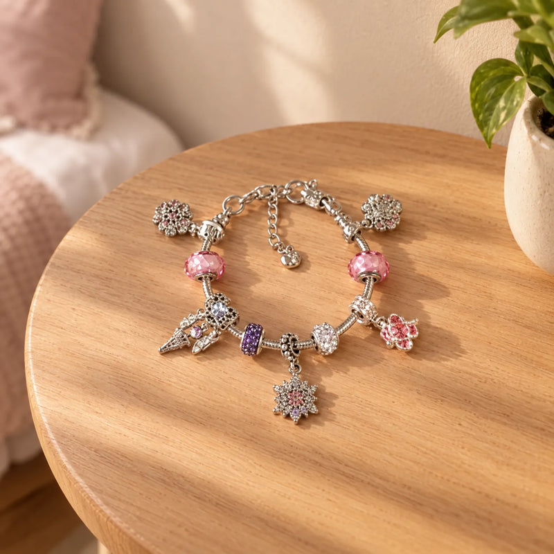 Afgewerkte zilverkleurige bedelarmband met roze, paarse en glinsterende bedels, liggend op een houten bijzettafel naast een plant. 1