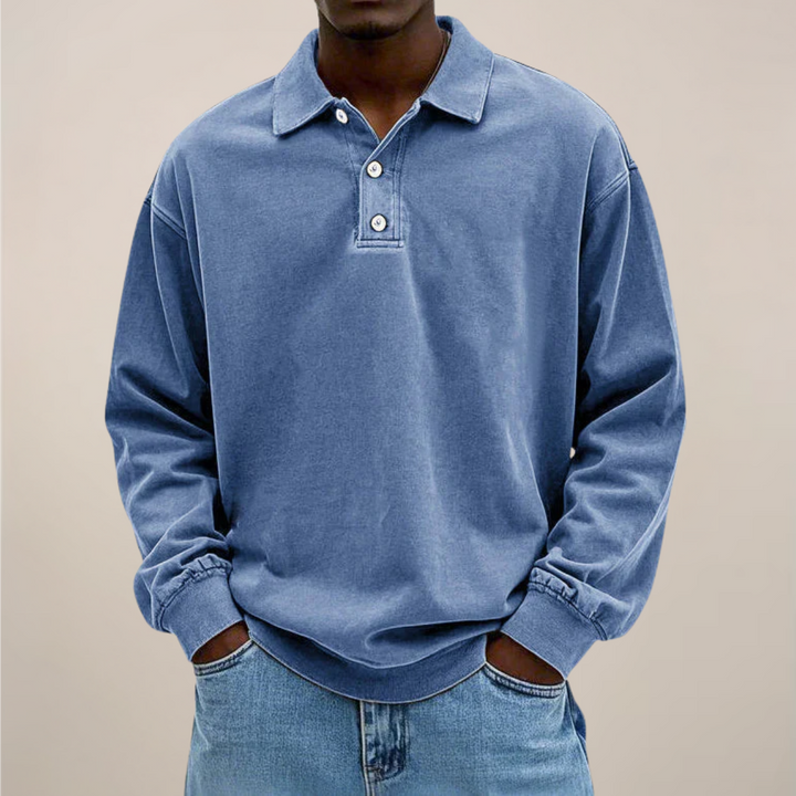 Model draagt de losse polo met lange mouwen en drie knopen in blauw, met handen in de zakken van een spijkerbroek. 1