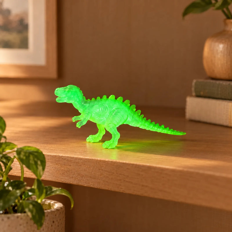 Doorschijnend groen dinosaurusfiguurtje met rugstekels, staand op een houten plank naast een plant en een stapel boeken. 3