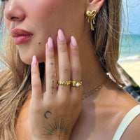 Close-up van een vrouw op het strand die een gouden smeltend hartoorbel draagt, met gouden ringen aan haar hand. 4
