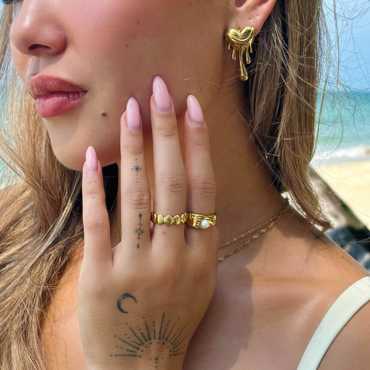 Close-up van een vrouw op het strand die een gouden smeltend hartoorbel draagt, met gouden ringen aan haar hand. 4