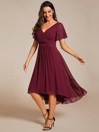 Model in bordeauxrode chiffon jurk met zwarte sandalen, kijkt lachend omlaag terwijl de asymmetrische rok zwiert. 47