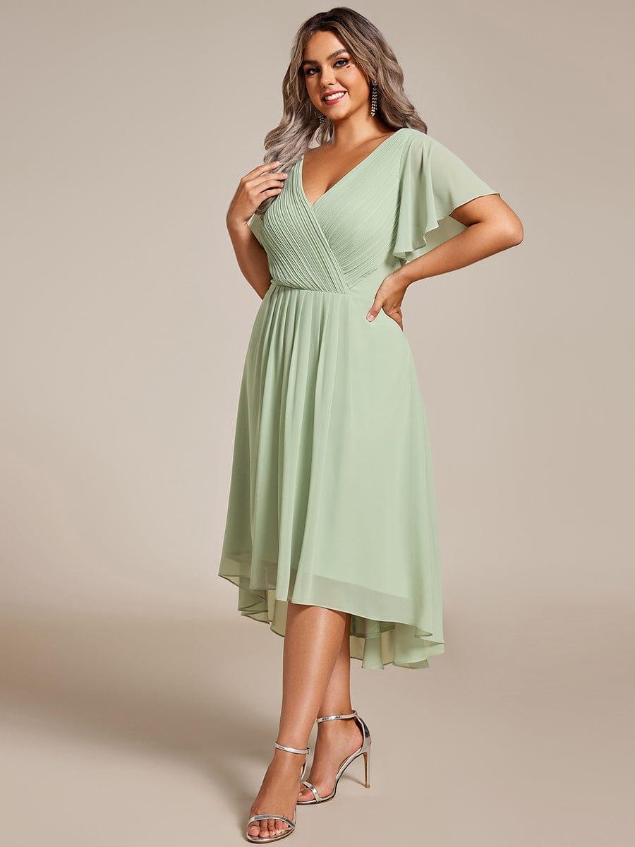 Model in saliegroene chiffon jurk met gekruiste geplooide V-hals en asymmetrische zoom, poseert met handen op de heup, zilveren sandalen. 81