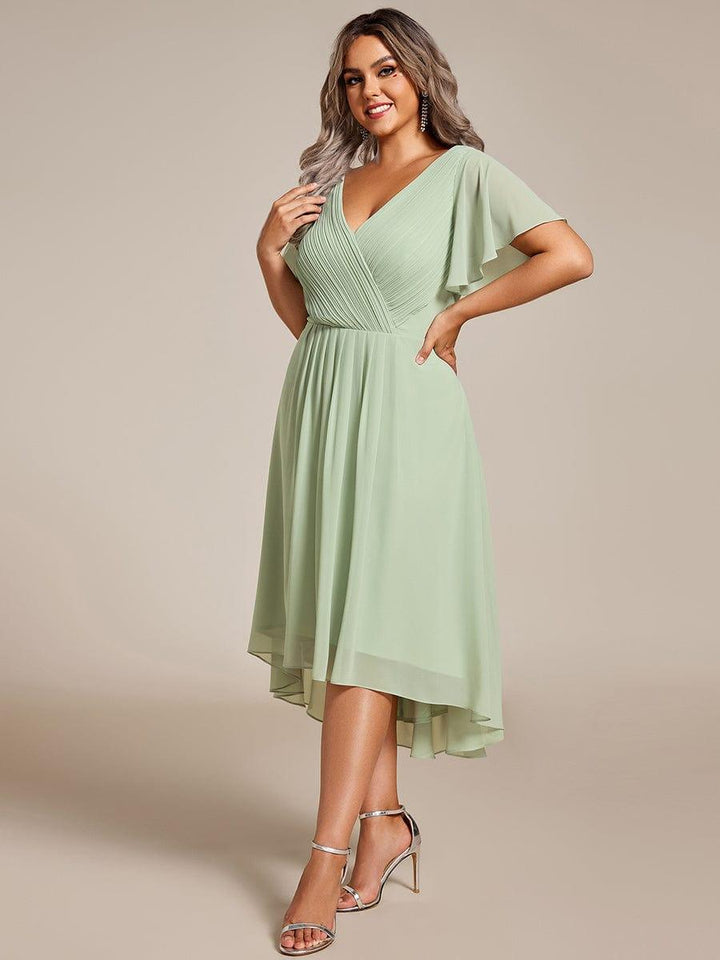Model in saliegroene chiffon jurk met gekruiste geplooide V-hals en asymmetrische zoom, poseert met handen op de heup, zilveren sandalen. 81