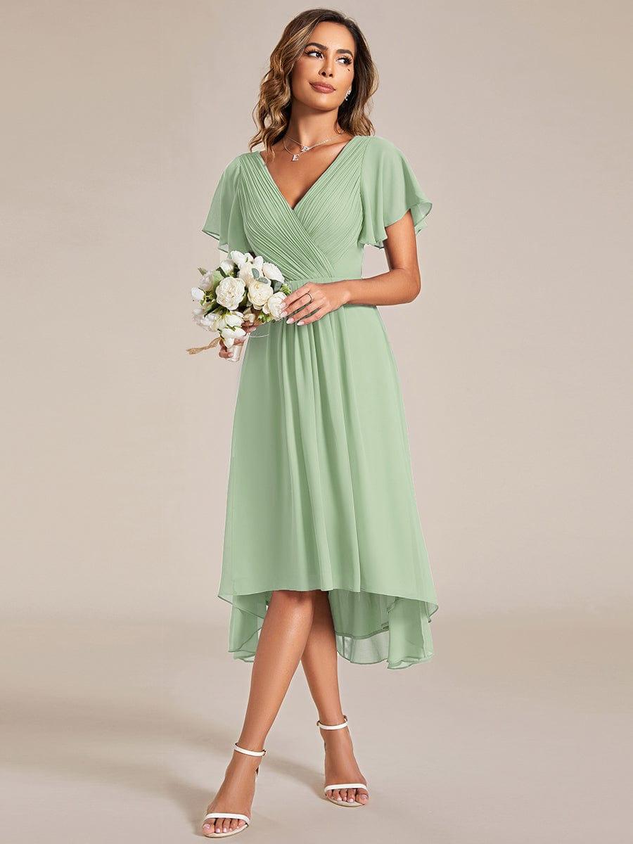 Model in saliegroene jurk houdt een klein boeket witte bloemen vast, met witte sandalen. 79
