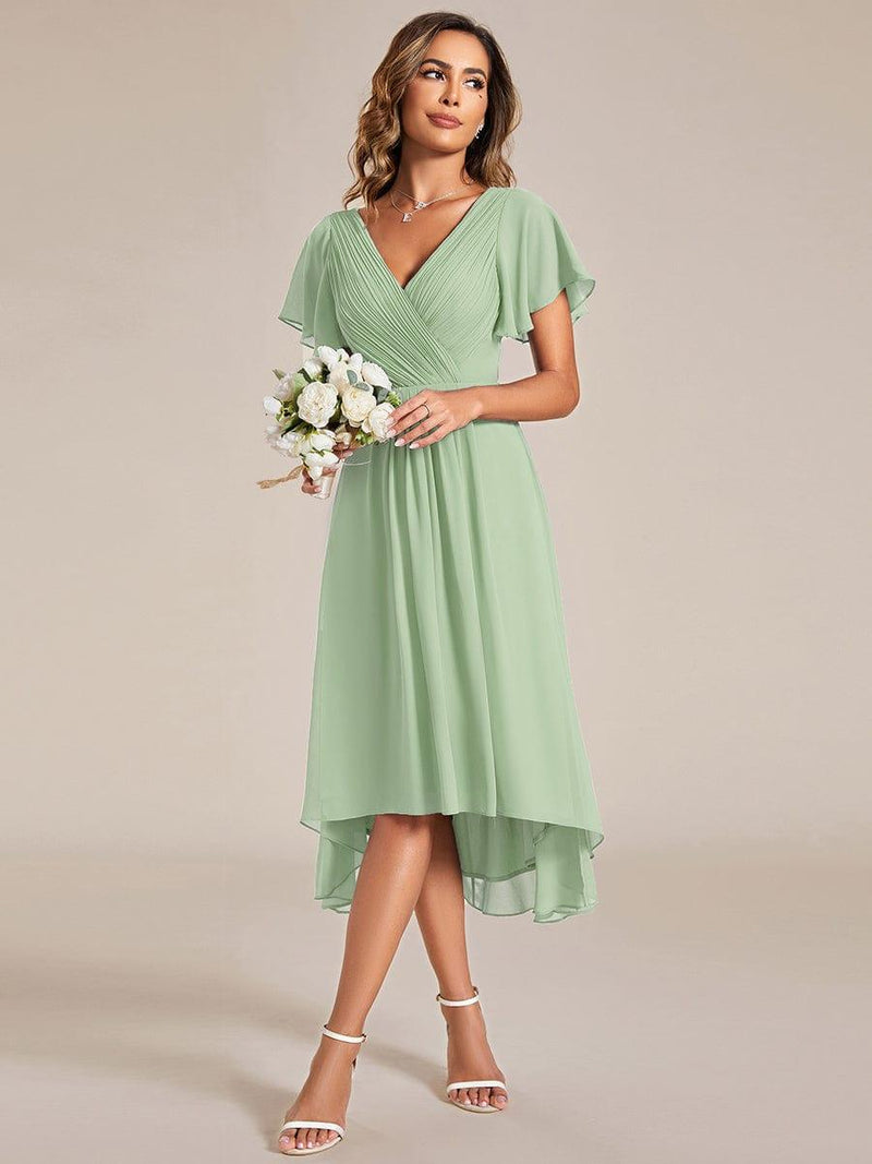 Model in saliegroene jurk houdt een klein boeket witte bloemen vast, met witte sandalen. 79