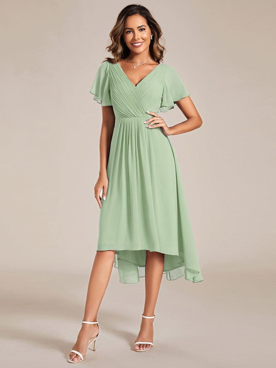Model in saliegroene jurk met asymmetrische zoom, hand op de heup, met witte sandalen en kettinghanger. 76