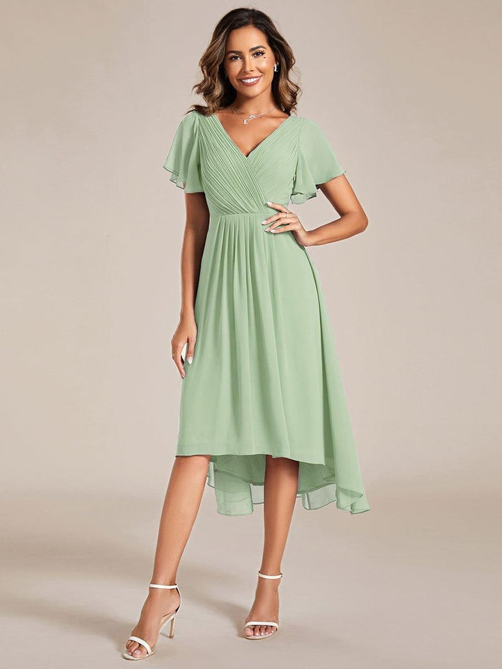 Model in saliegroene jurk met asymmetrische zoom, hand op de heup, met witte sandalen en kettinghanger. 76