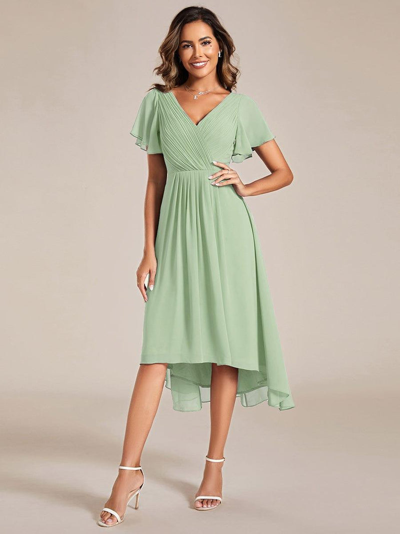 Model in saliegroene jurk met asymmetrische zoom, hand op de heup, met witte sandalen en kettinghanger. 76