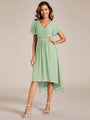 Model in saliegroene jurk met asymmetrische zoom, hand op de heup, met witte sandalen en kettinghanger. 76