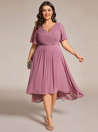 Model met maatje meer draagt de oudroze jurk met asymmetrische zoom, met zilverkleurige clutch en sandalen, frontaal gefotografeerd. 63