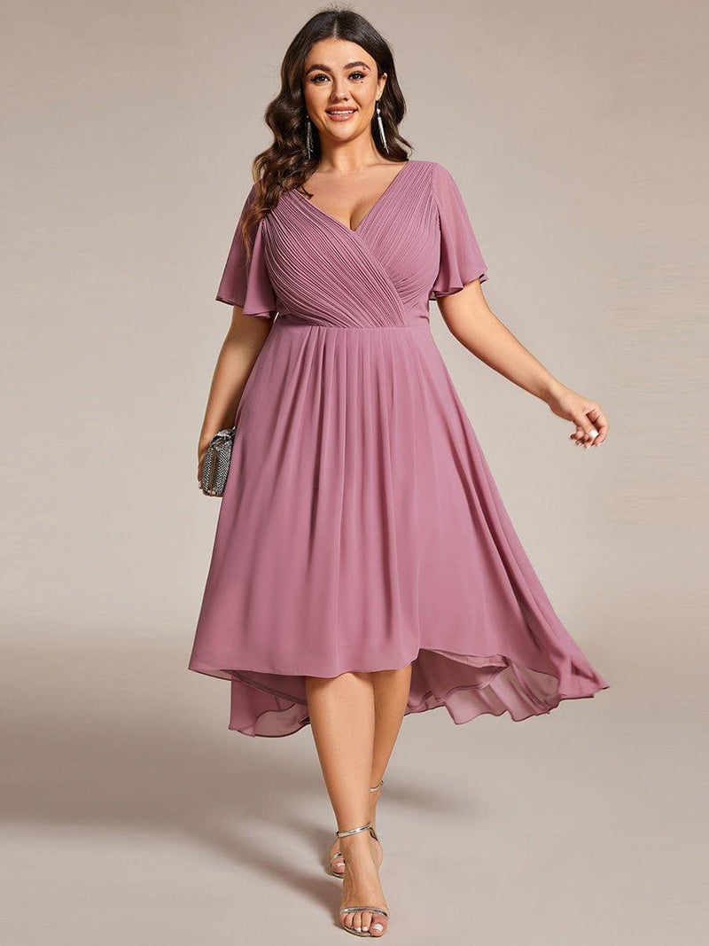 Model met maatje meer draagt de oudroze jurk met asymmetrische zoom, met zilverkleurige clutch en sandalen, frontaal gefotografeerd. 63